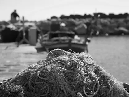 Primo piano di reti da pesca ammassate su una banchina, con barche e una figura sfocati dietro, in bianco e nero