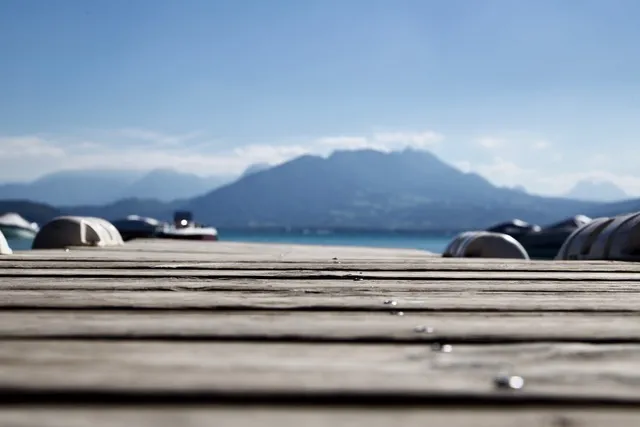 Assi di un pontile di legno in primo piano con lago, barche e montagne sfocati sullo sfondo sotto il cielo azzurro