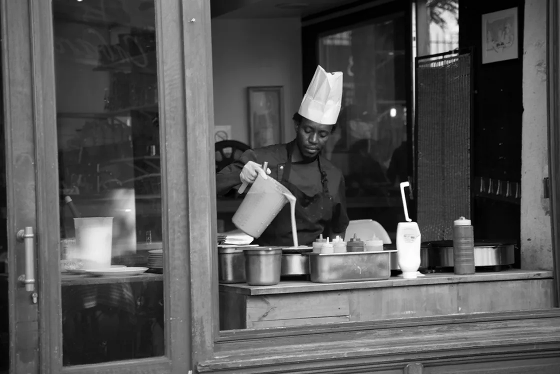 Cuoco con cappello di carta versa una pastella da una brocca, visto attraverso la vetrina di un locale, in bianco e nero