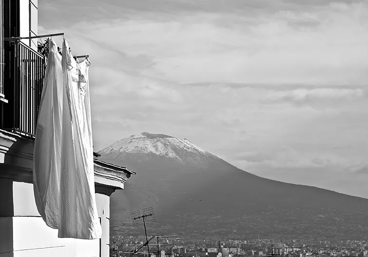 Panni bianchi stesi a un balcone con la cima innevata del Vesuvio sullo sfondo, in bianco e nero
