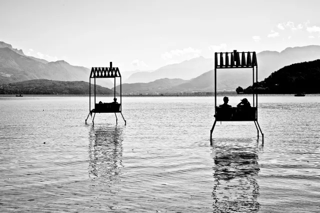 Due strutture con tettoia immerse nell'acqua del lago con persone sedute e montagne sullo sfondo, in bianco e nero