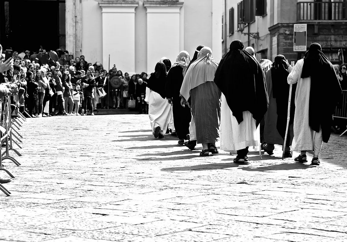 Una processione di figure incappucciate e in tunica attraversa una piazza lastricata davanti alla folla, in bianco e nero