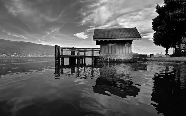 Casetta di legno sul lago accanto a un pontile, con riflessi nell'acqua e un albero sulla destra, in bianco e nero