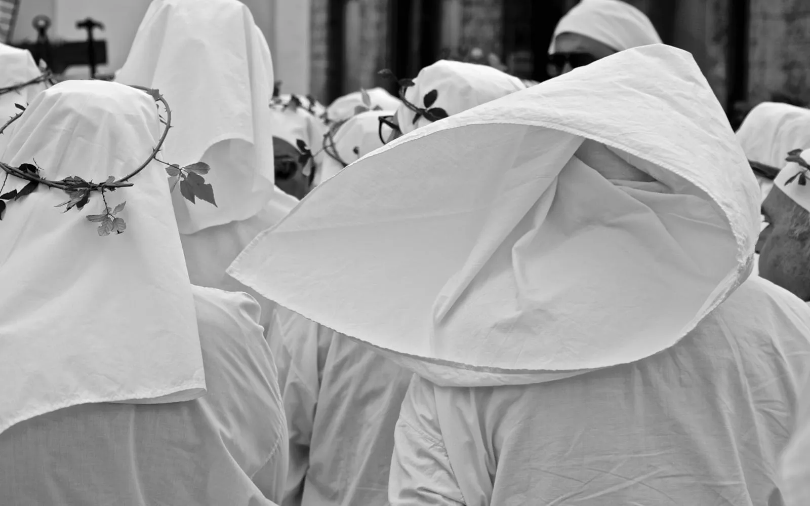 Penitenti incappucciati di bianco, alcuni con corone di spine e foglie sul capo, ripresi da vicino, in bianco e nero