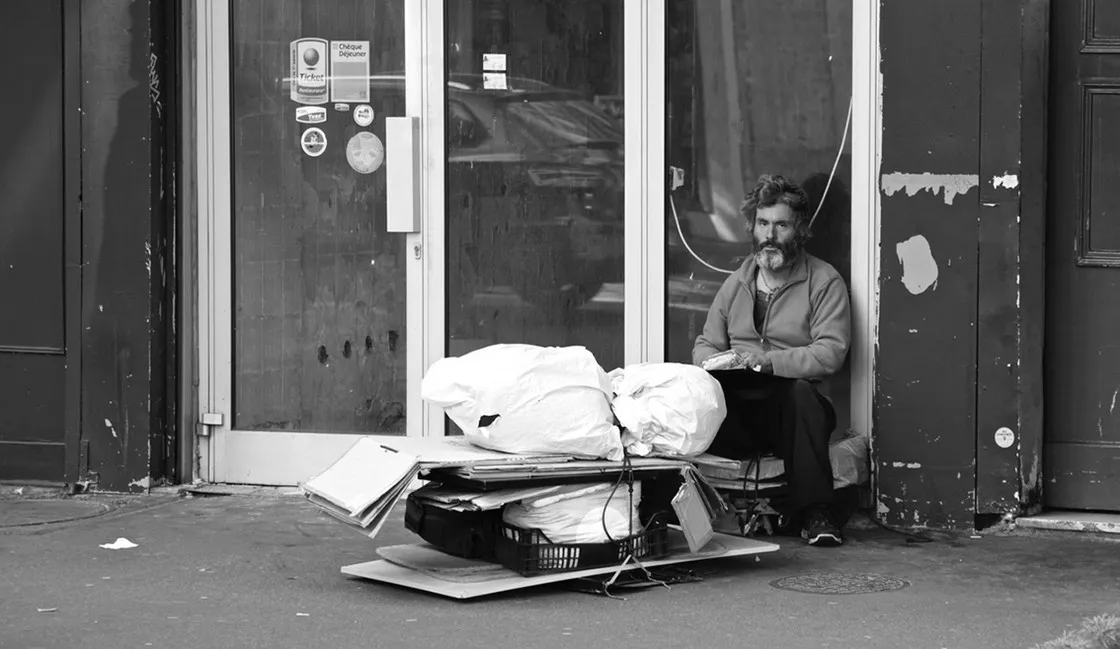 Uomo senza dimora seduto nel vano di una porta con i suoi averi ammassati sul marciapiede, in bianco e nero