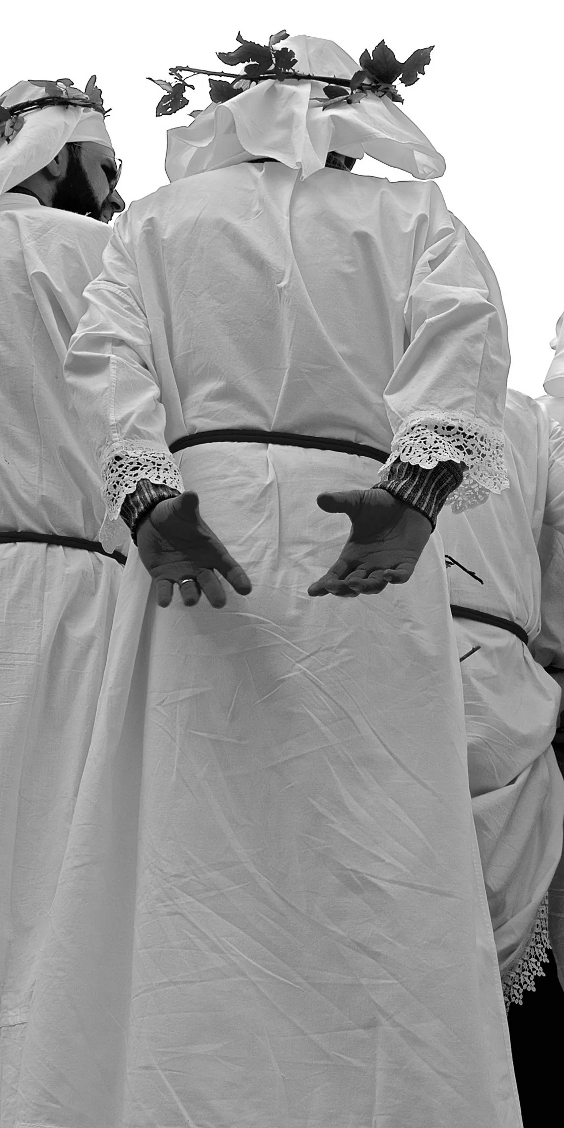Figure in tunica bianca con polsini di pizzo e corone di foglie, riprese dal basso con le mani aperte, in bianco e nero