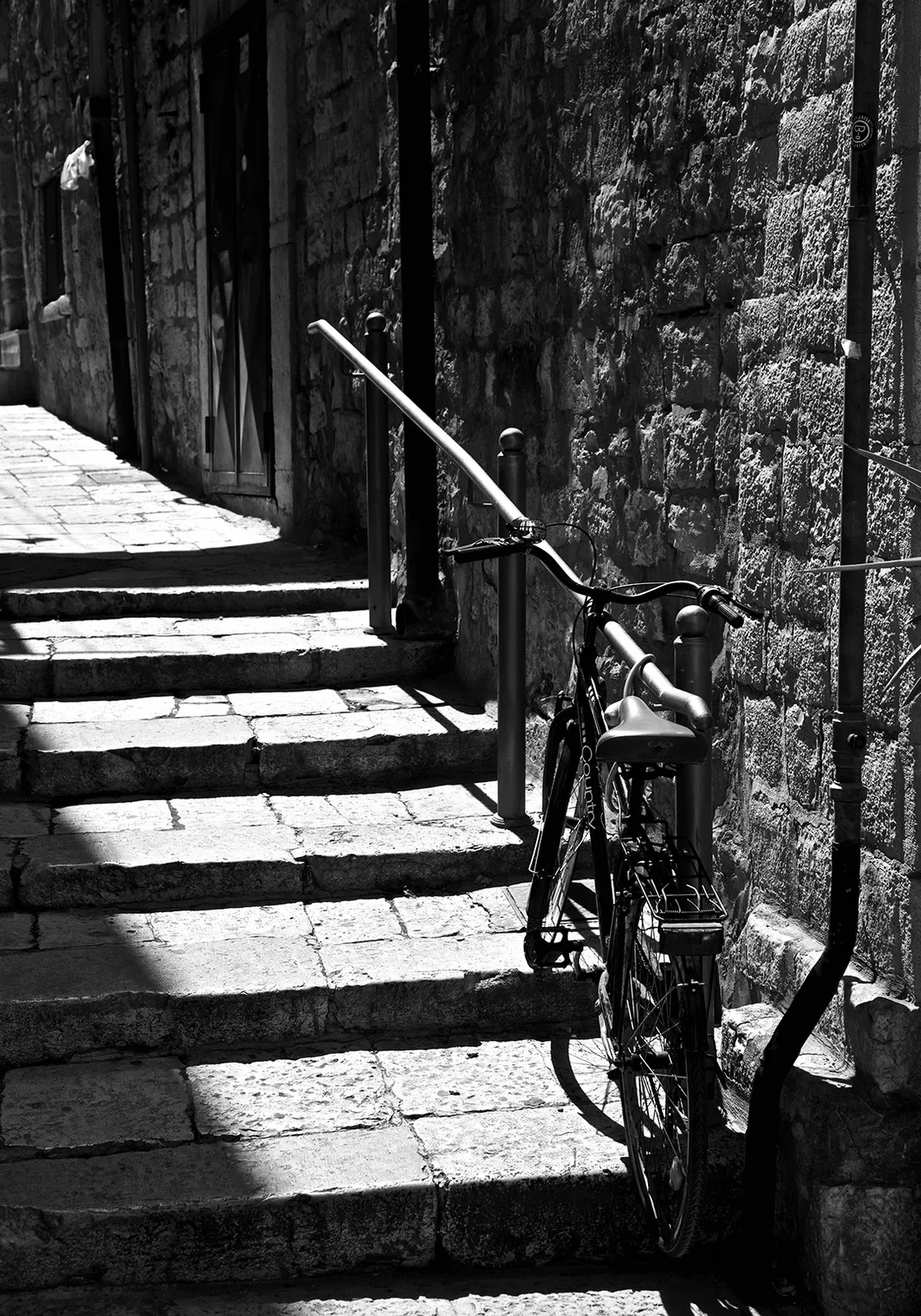 Una bicicletta appoggiata a un muro di pietra lungo la scalinata di un vicolo assolato, in bianco e nero