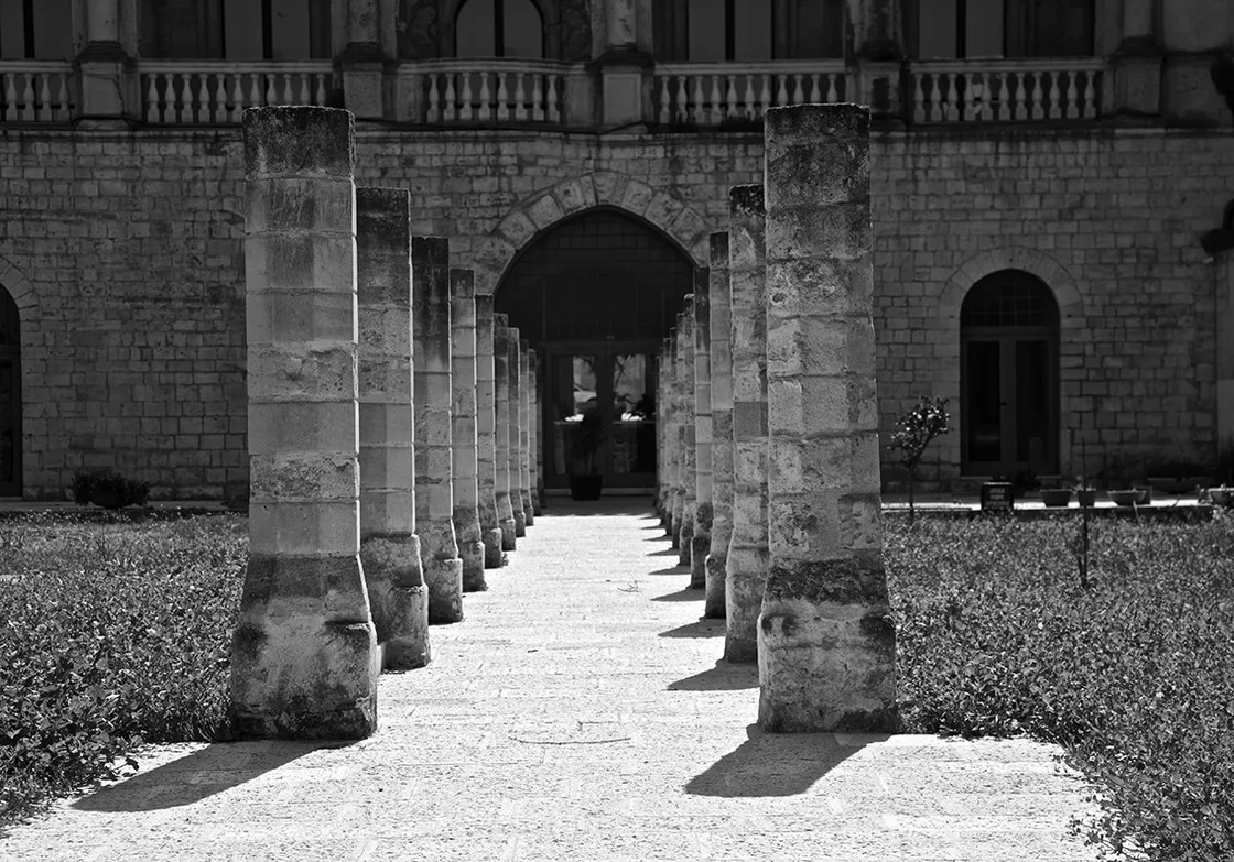 Doppia fila di pilastri di pietra tronchi lungo un vialetto tra siepi, verso l'ingresso ad arco di un edificio, in bianco e nero