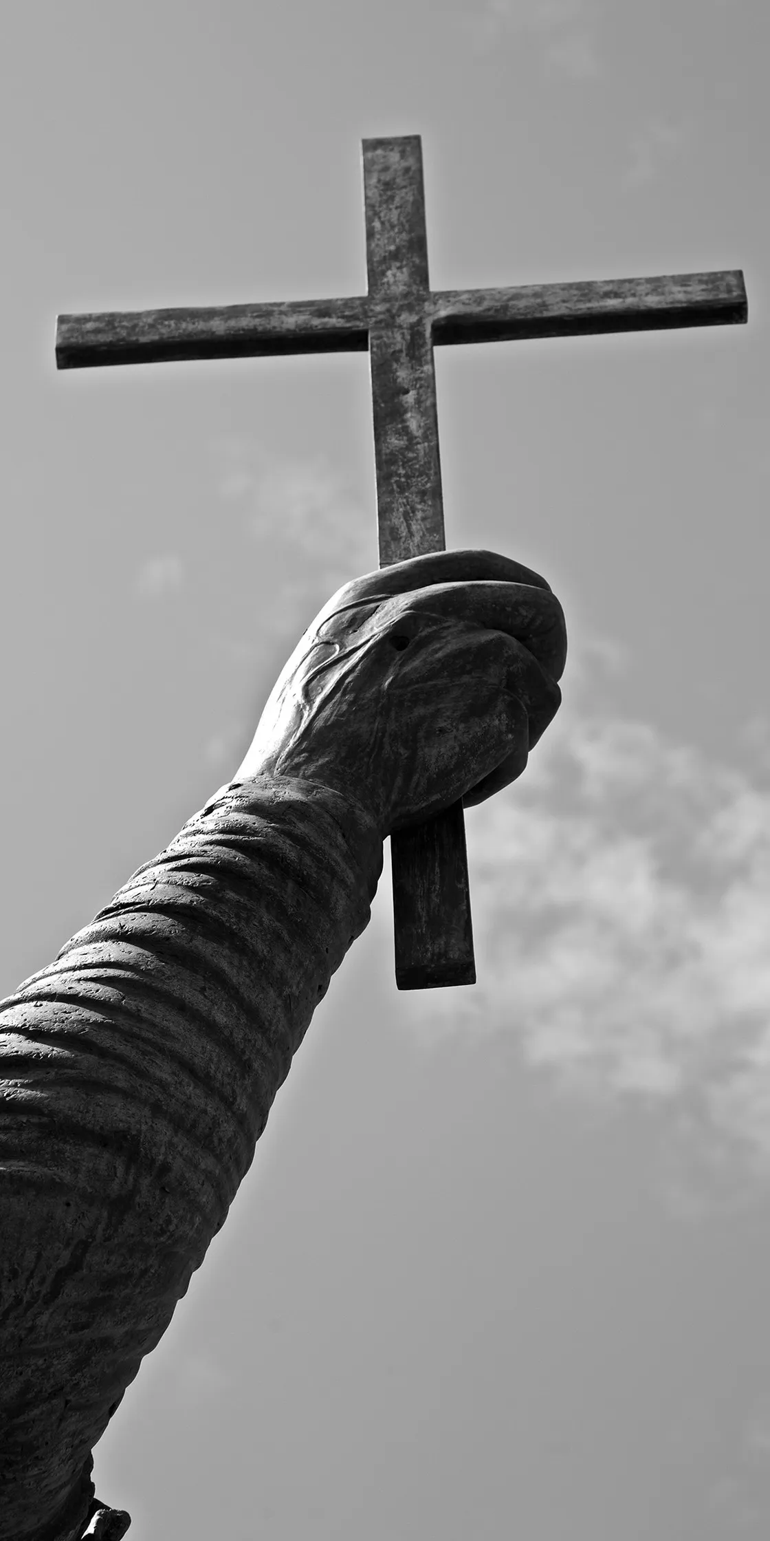Il braccio di una statua che regge in alto una croce, stagliato contro il cielo, in bianco e nero