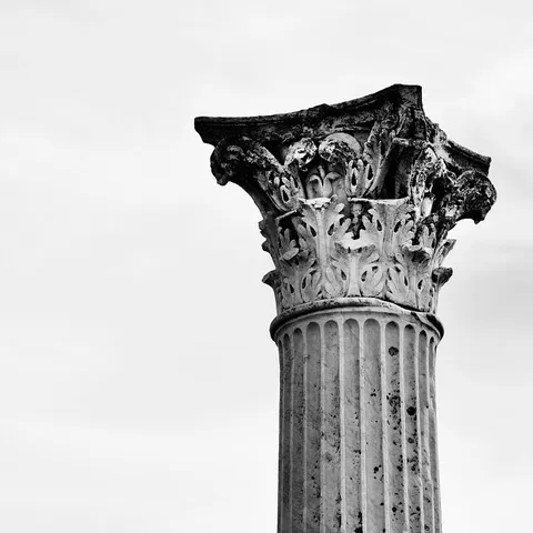 Capitello corinzio in cima al fusto scanalato di un'antica colonna, contro un cielo chiaro, in bianco e nero