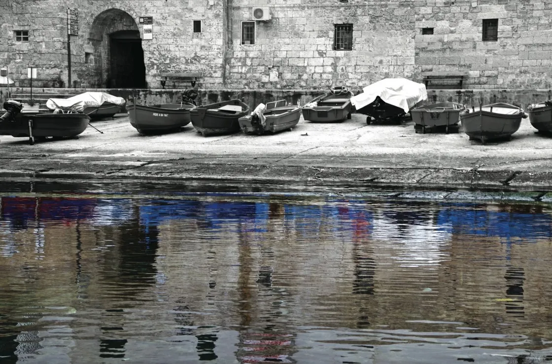 Barche in secca su una banchina davanti a un muro di pietra, con riflessi colorati che vibrano nell'acqua