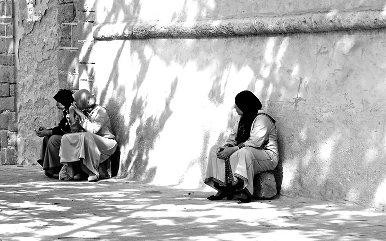 Donne con il velo sedute contro un muro assolato screziato di ombre, in bianco e nero