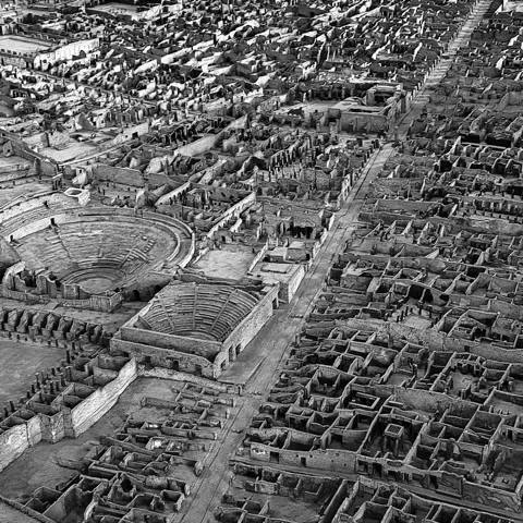 Veduta aerea di un sito archeologico con un anfiteatro e la griglia di rovine di un'antica città, in bianco e nero