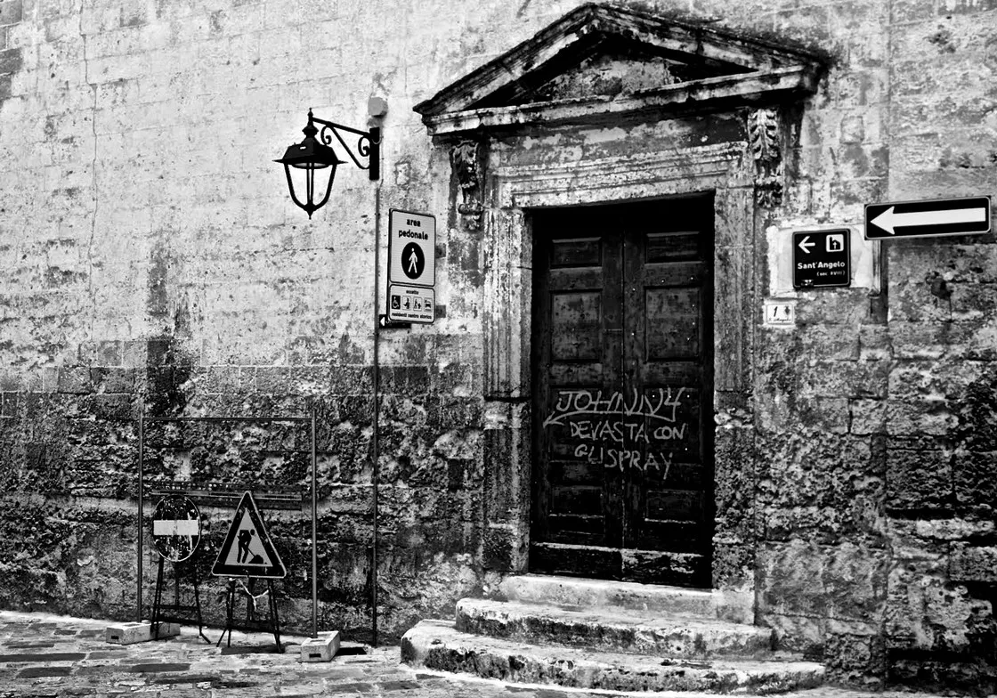 Un portale antico con timpano e porta di legno segnata da graffiti, tra cartelli stradali e un lampione, in bianco e nero