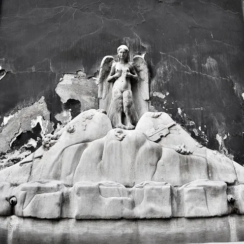 Scultura in marmo di una figura alata su un cumulo roccioso, davanti a una parete scrostata, in bianco e nero