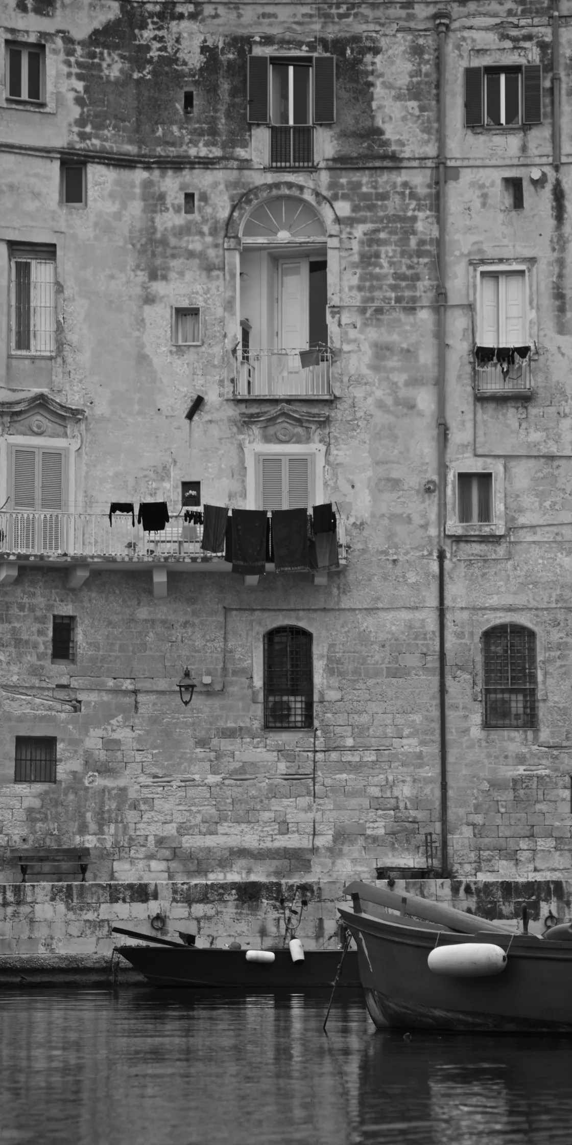 Alta facciata in pietra di un antico edificio sul porto, con panni ai balconi e barche ormeggiate alla base, in bianco e nero