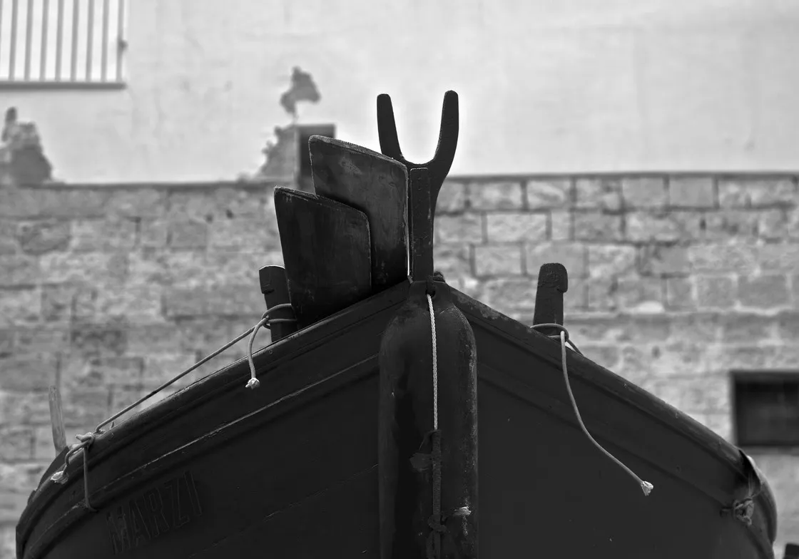 Dettaglio della prua di una barca in secca con scalmo di legno a forcella, davanti a un muro di pietra, in bianco e nero