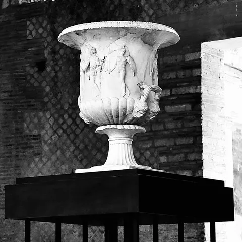 Grande vaso di marmo con figure a rilievo, posato su un piedistallo scuro davanti a un muro di mattoni, in bianco e nero