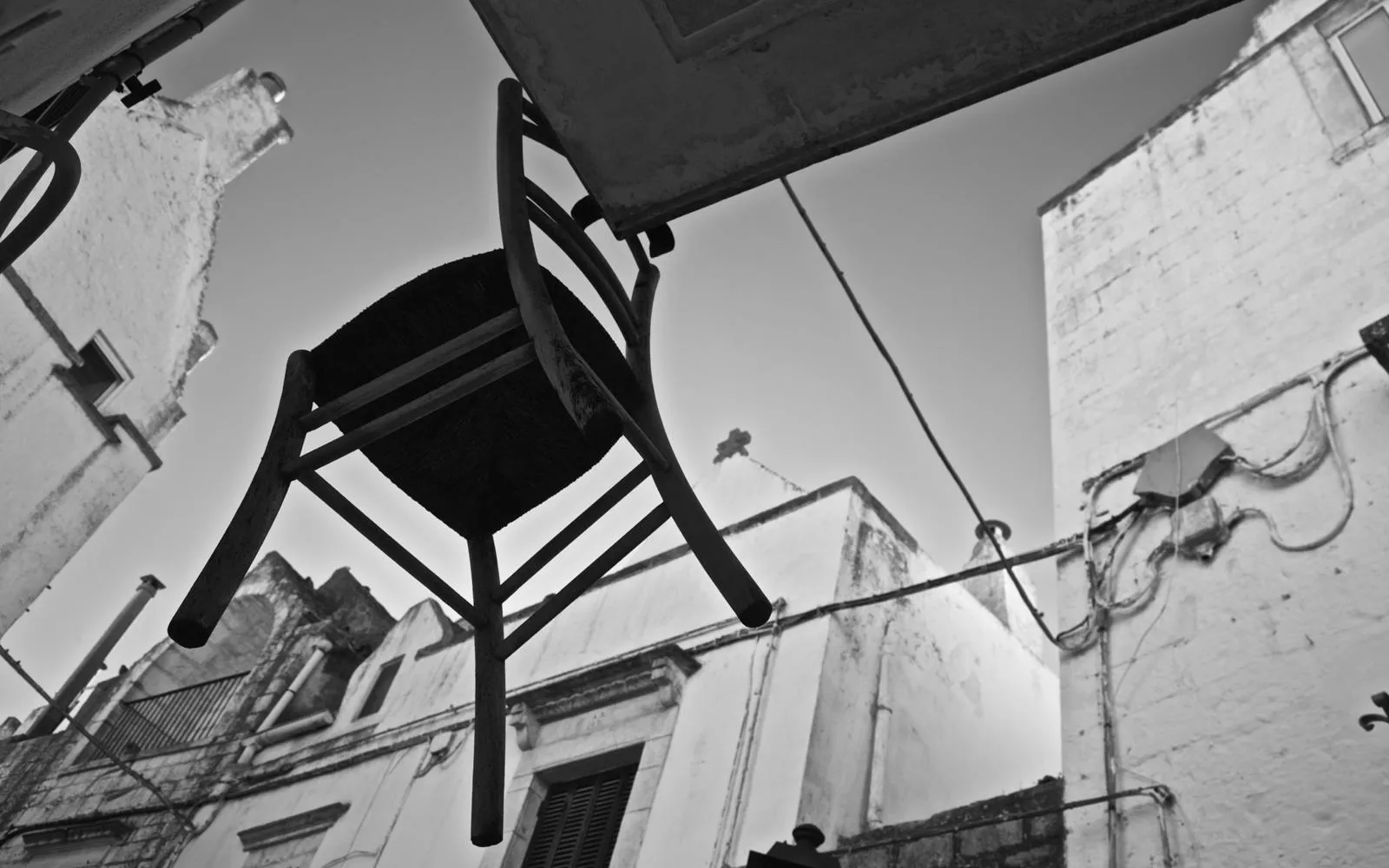 Una sedia impagliata capovolta in alto tra i muri di vecchie case, vista dal basso contro il cielo, in bianco e nero