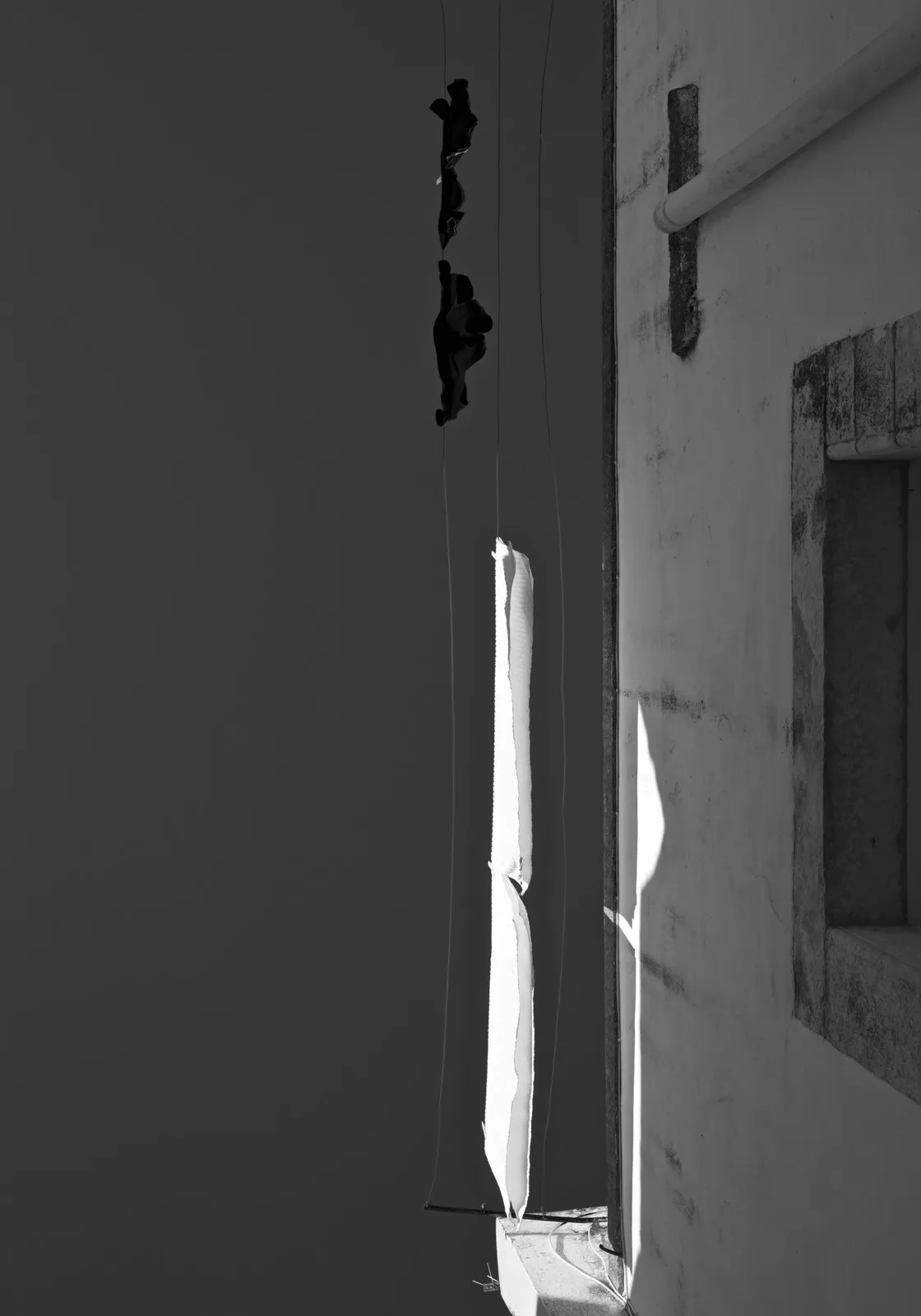 Panni scuri e un telo bianco stesi a fili sottili all'angolo di un edificio, tra luce e ombra nette, in bianco e nero