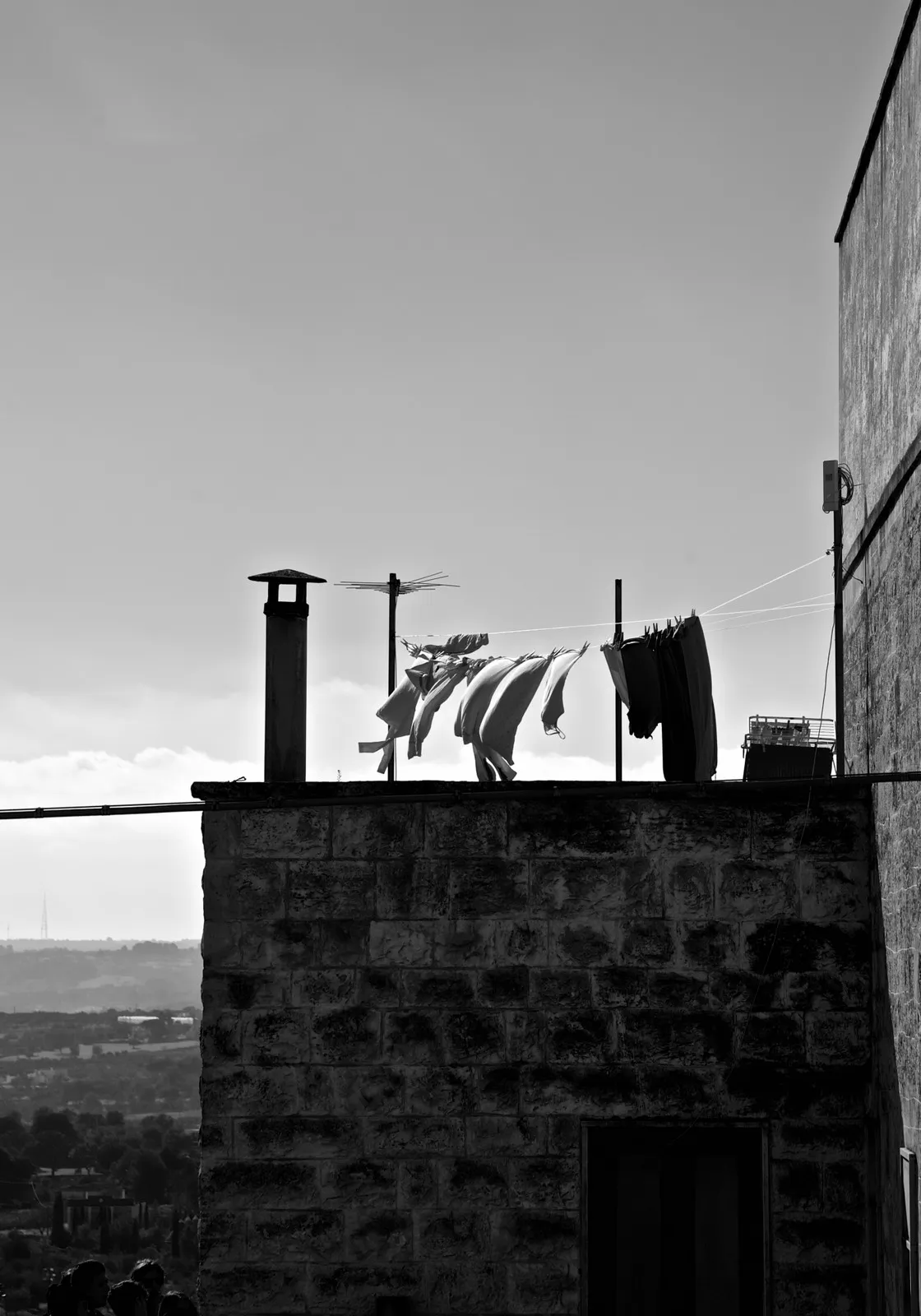 Panni stesi al vento sulla terrazza di un edificio in pietra, con un ampio paesaggio collinare sullo sfondo, in bianco e nero