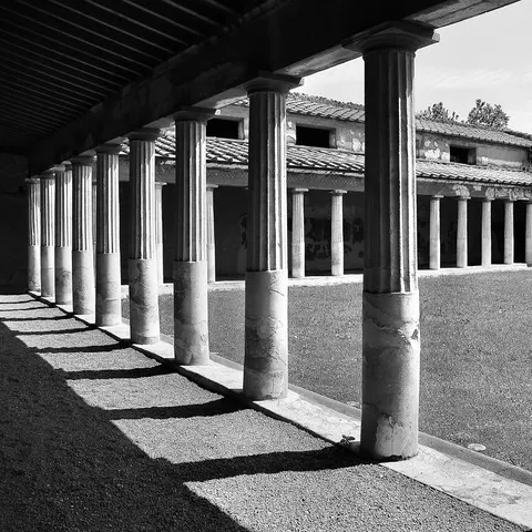 Colonnato di un'antica villa con le colonne che proiettano ombre sul terreno e un cortile sullo sfondo, in bianco e nero