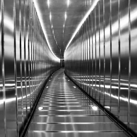 Tunnel metallico moderno con pareti in acciaio scanalato e luci a terra che convergono in prospettiva, in bianco e nero