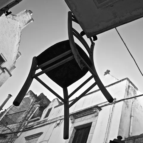 Sedia impagliata ripresa dal basso contro il cielo, con i vecchi edifici di un paese sullo sfondo, in bianco e nero