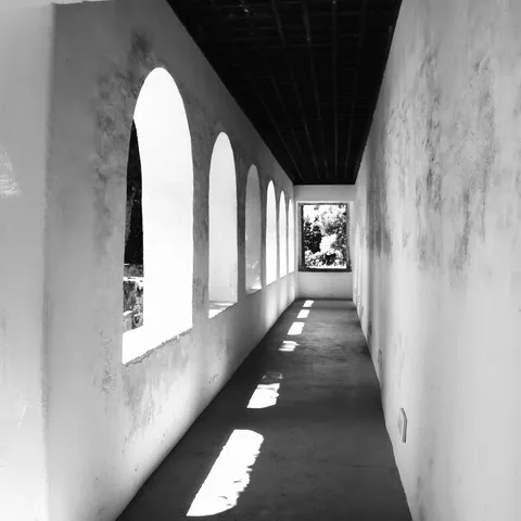 Corridoio imbiancato con una fila di aperture ad arco che lasciano entrare la luce sul pavimento, in bianco e nero