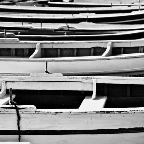 Diverse barche a remi bianche ormeggiate affiancate, con gli scafi e le panche che si sovrappongono, in bianco e nero