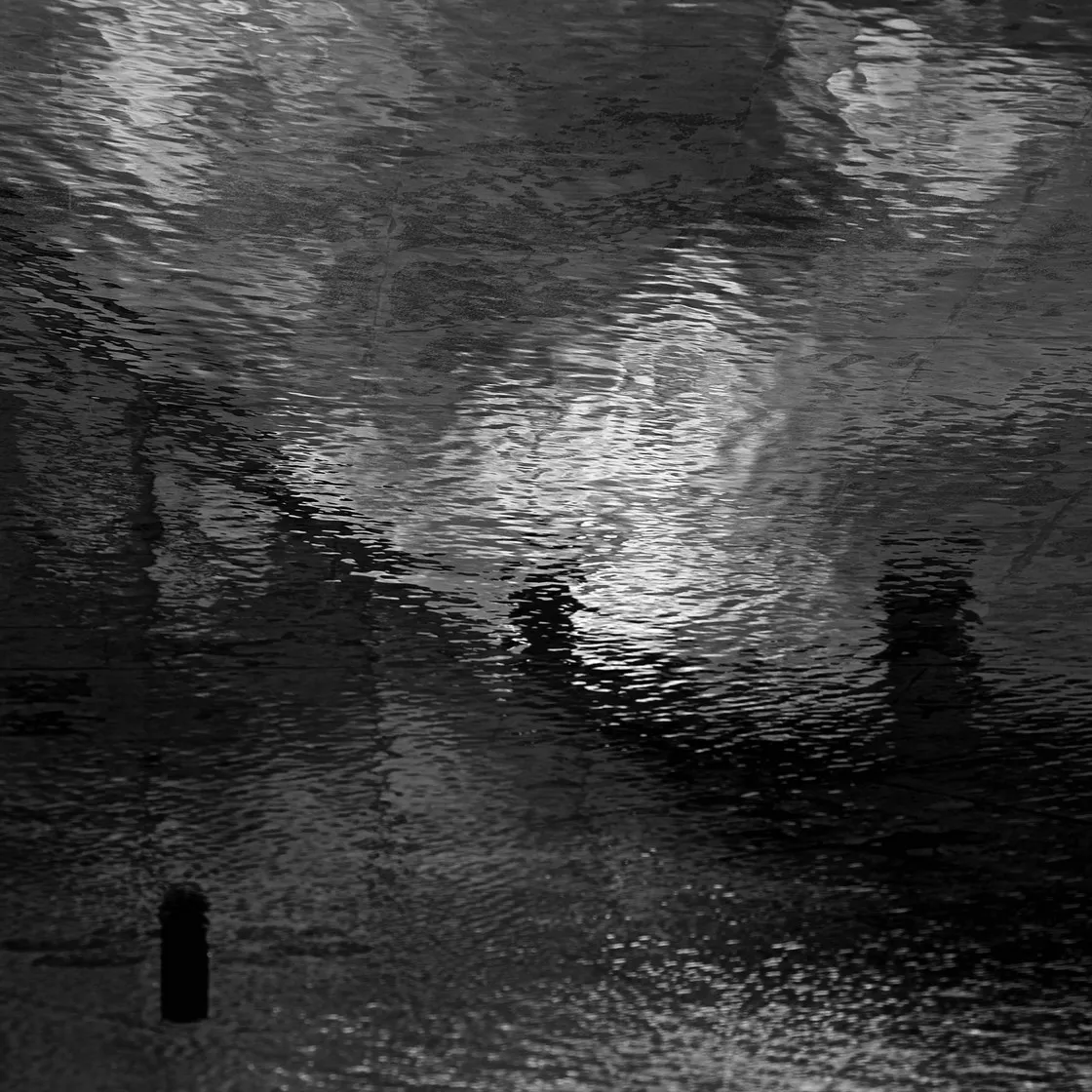 Superficie d'acqua scura e increspata con riflessi di luce e un piccolo palo affiorante in basso, in bianco e nero