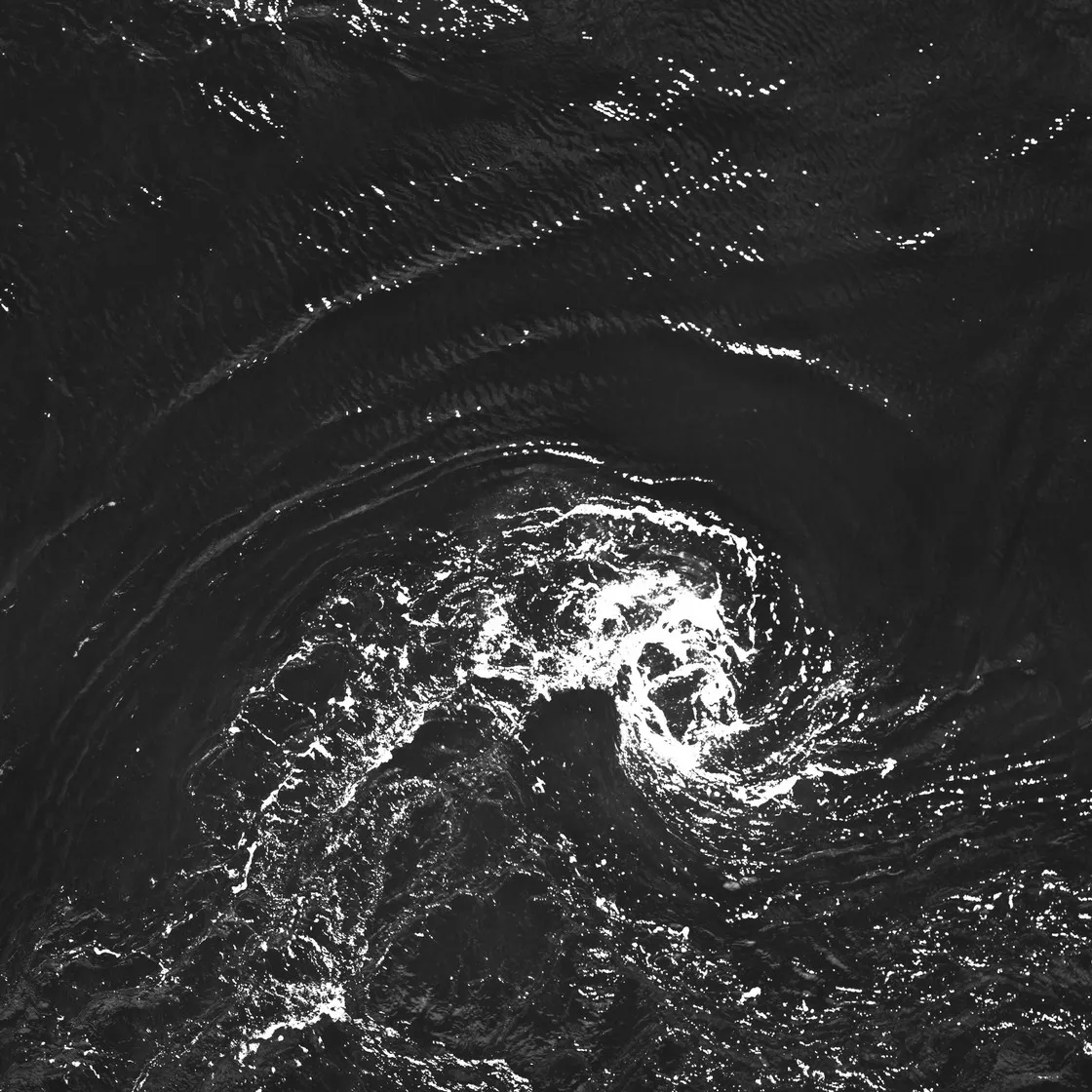 Vortice d'acqua ripreso dall'alto, con spuma bianca che spiraleggia sulla superficie scura del mare, in bianco e nero