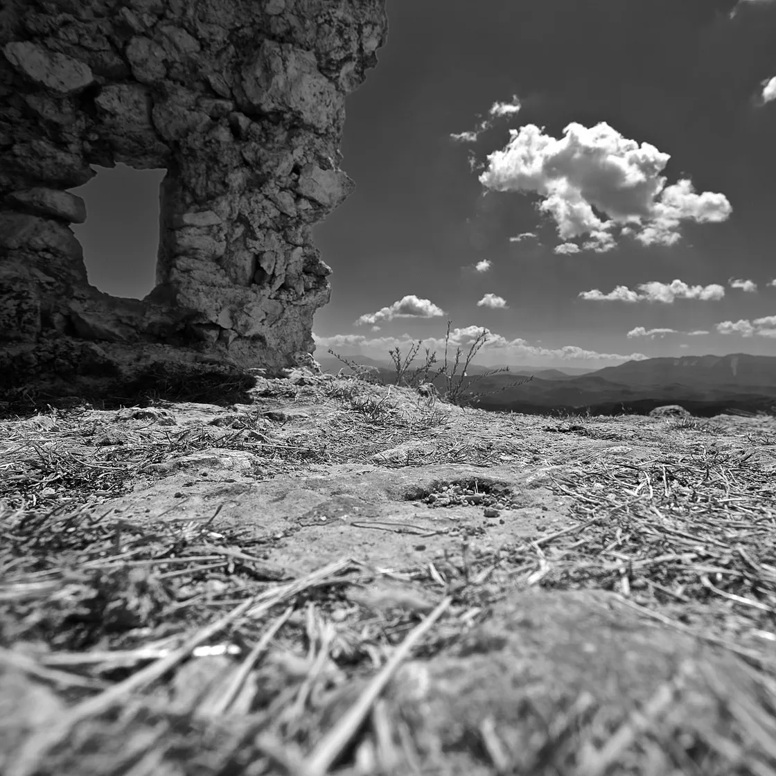 Muro diroccato in pietra con un'apertura, ripreso dal suolo tra erba secca, con nuvole e monti lontani, in bianco e nero