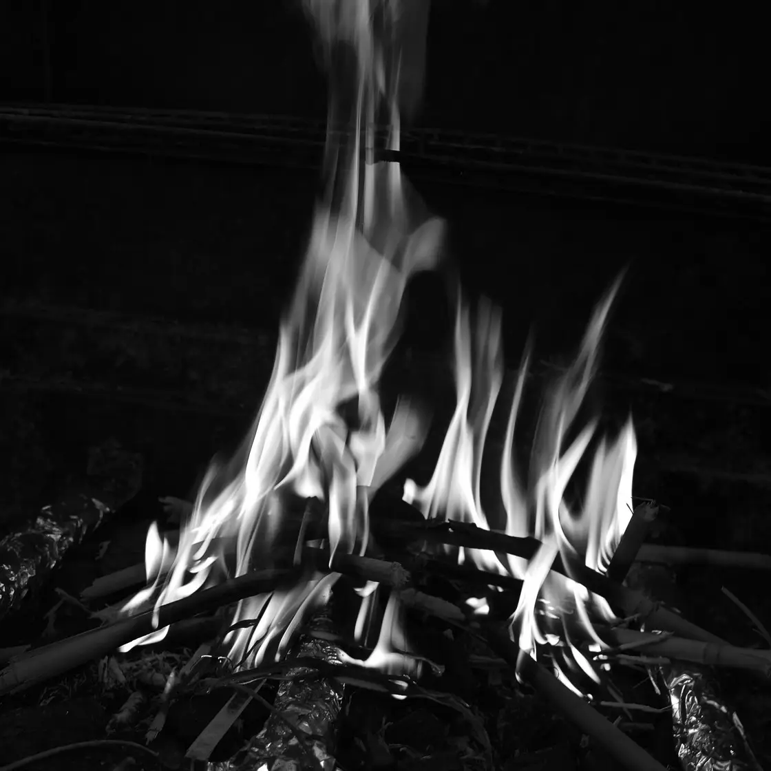 Fiamme che si levano da un fuoco di ramoscelli su uno sfondo scuro, in bianco e nero
