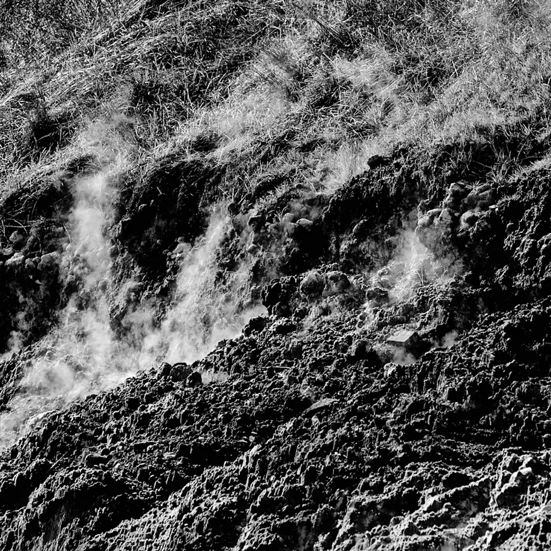 Sbuffi di fumo bianco che salgono da un pendio roccioso e sassoso con erba secca in alto, in bianco e nero